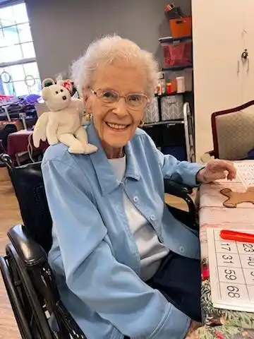 Smiling older woman seated at an activity table with a stuffed animal on her shoulder.